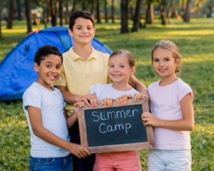 Four children at summer camp holding a chalkboard sign that says “Summer Camp” with camping tents and trees in the background.