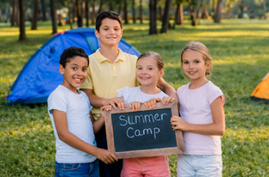 Four children at summer camp holding a chalkboard sign that says “Summer Camp” with camping tents and trees in the background.