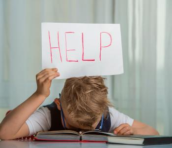 A child with his head down on a book, holding a "help" sign